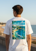 Person wearing a white t-shirt with a scenic design of Biarritz on the back, standing by the ocean.